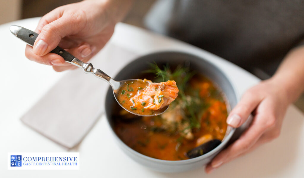 Person holding a spoon of fish soup with herbs in a gray bowl.